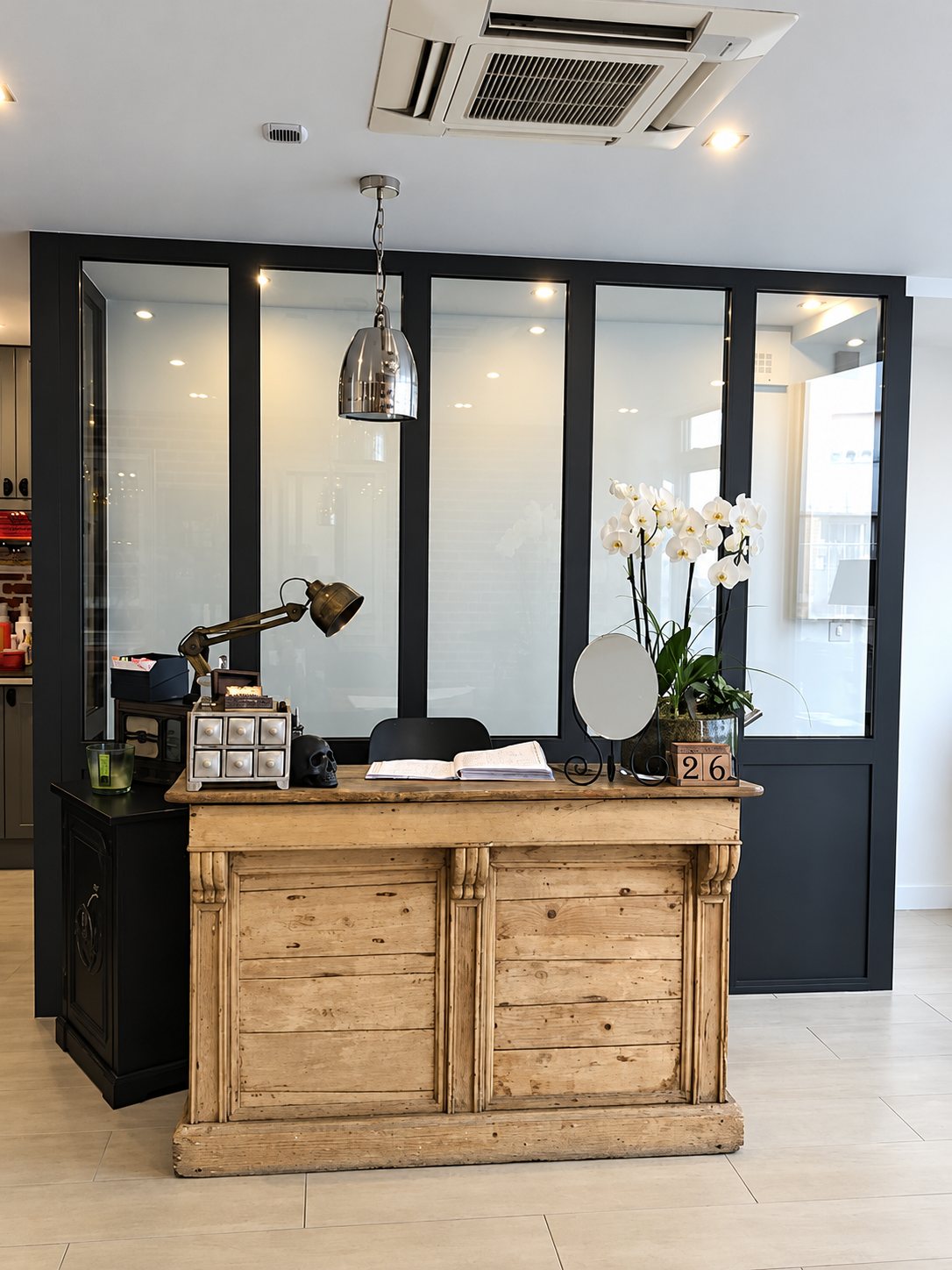 Comptoir d’accueil en vieux bois, lampe articulée, miroir et orchidée blanche devant la verrière noire du salon.