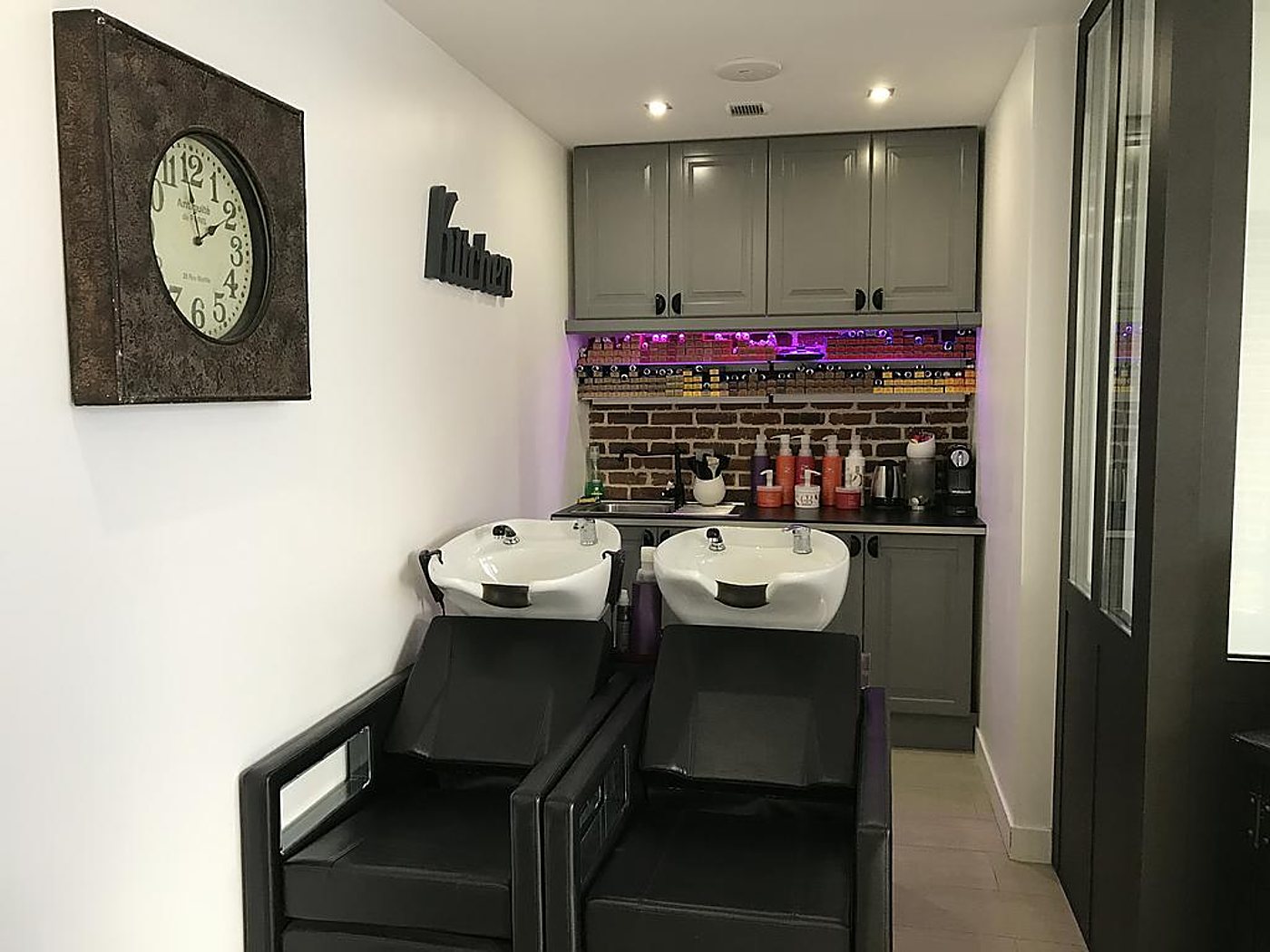Espace de lavage du salon avec deux bacs de shampoing blancs, une horloge murale et des fauteuils noirs.
