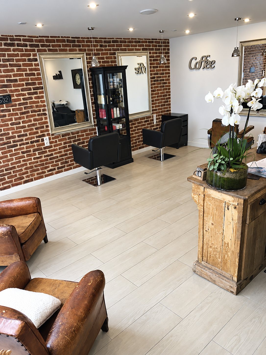 Salon vu depuis les fauteuils en cuir vieilli : mur de briques, miroirs et fauteuils de coiffeur noirs.